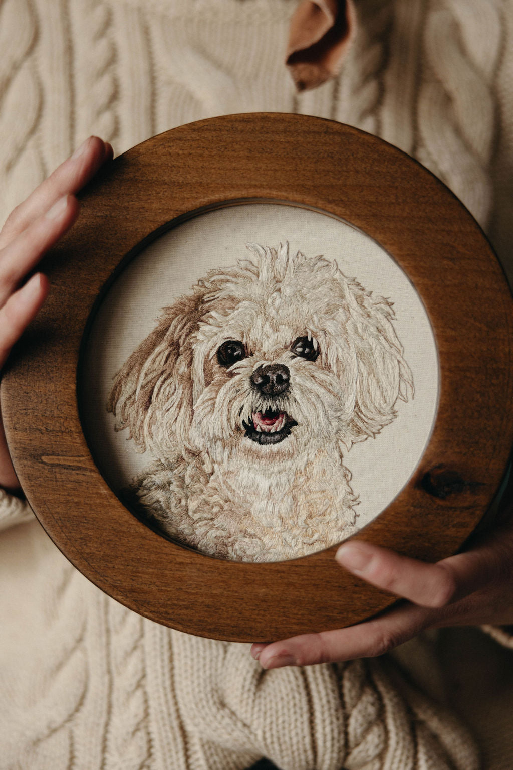 Embroidered portrait of a small white curly haired dog in a round walnut frame held by caucasian hands against a knitted white sweater